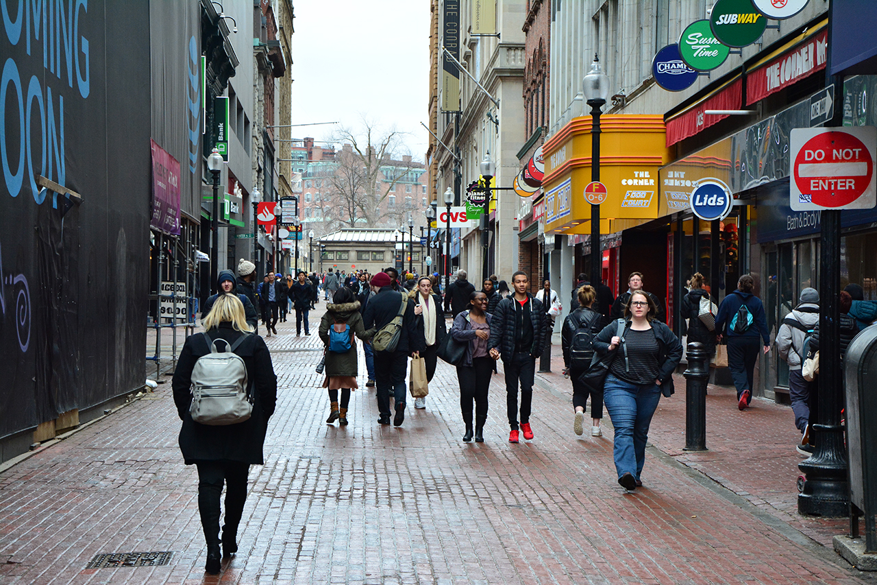 People walk in multiple directions down a narrow cobblestone street in Downtown Crossing, past rows of shops. 