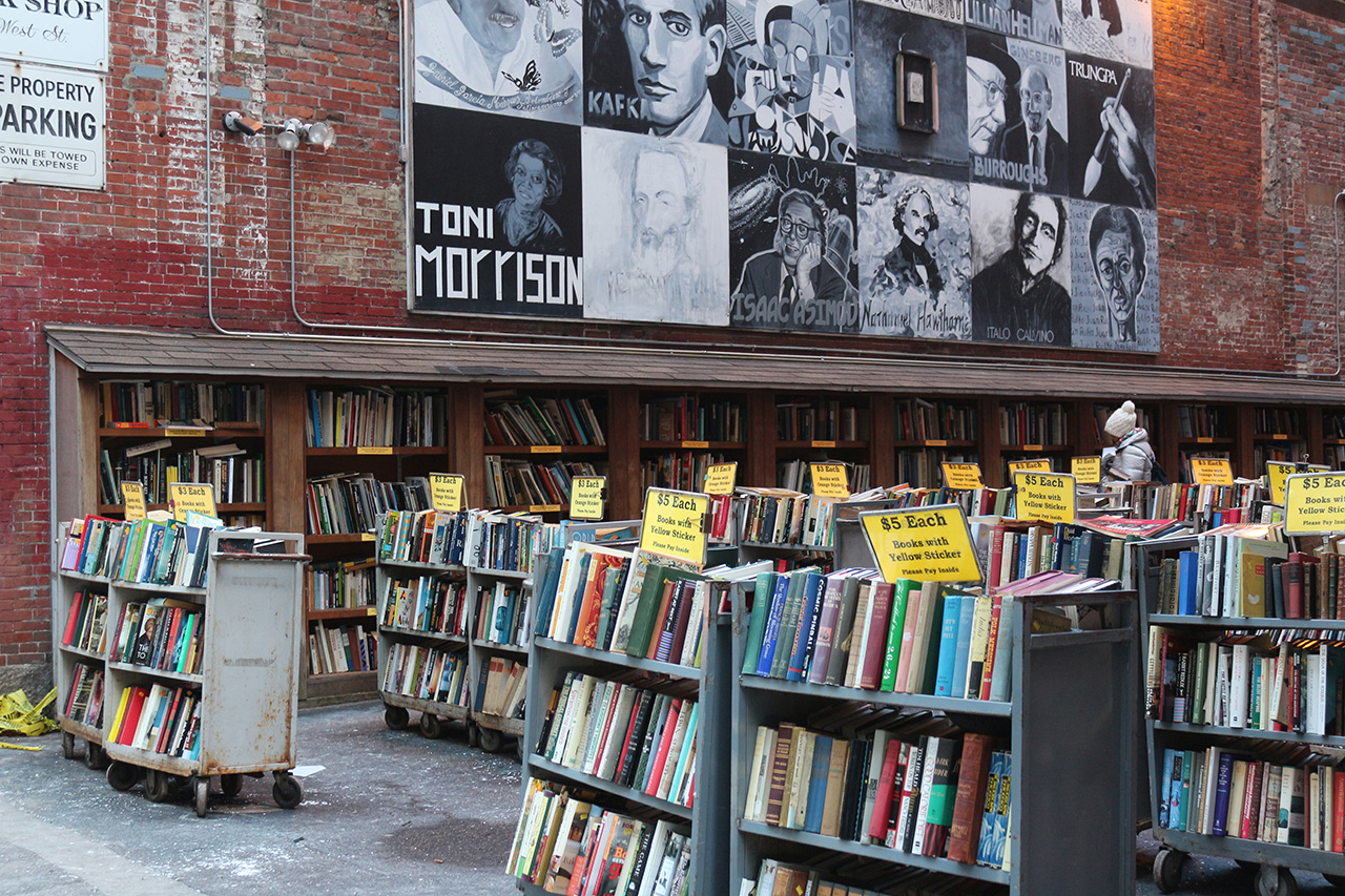 This image shows the open lot beside Brattle Book Shop which is full of carts of books. The brick wall of the book shop is painted with a mural of different authors.