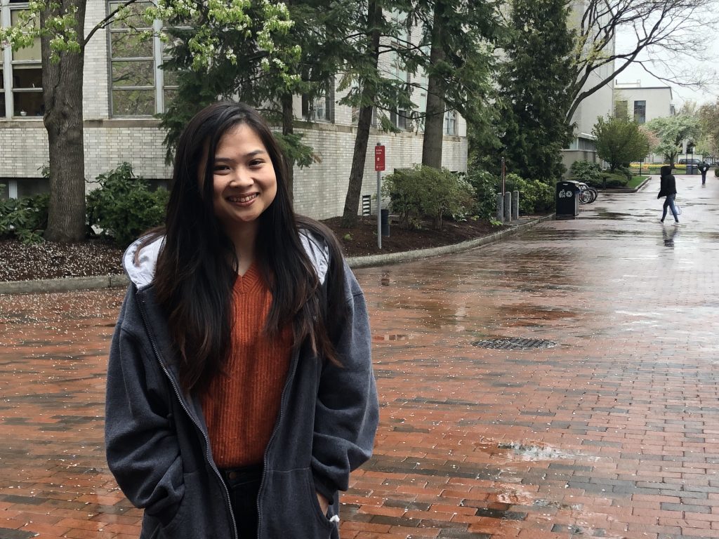 Nesanne Mae Tam, a college sophomore, stands on the brick walkway outside Northeastern's library. She has long black hair and is wearing a grey sweatshirt. 