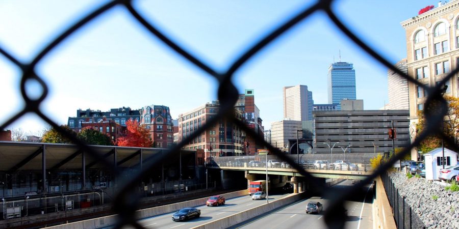 this photo shows boston highway and skyline visible through a chain link fence