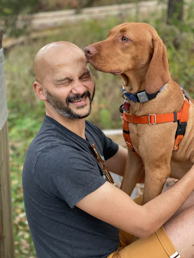 A man with a shaved head and beard sits with his dog on his lap. His eyes are closed as the dog’s nose presses lightly against his forehead. The dog is wearing an orange harness and has tan fur.