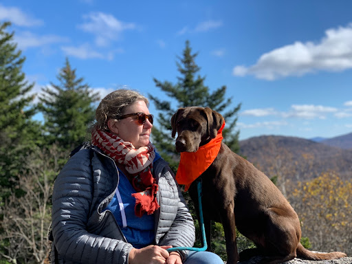 A white woman with light hair is bundled up in winter layers and sunglasses. Beside her is a brown labrador wearing a red bandana. They are seated in front of a blue sky and a forest filled with trees.
