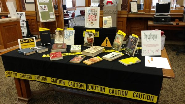 Banned books are displayed on a black table in a library. The table is wrapped in caution tape to symbolize readers' lack of access to them. 