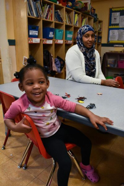 A young girl with dark skin and in a pink long-sleeve shirt smiles while sitting at a blue table in a red chair. An adult —the young girl's teacher — wearing a white top and dark blue headscarf, sits on the other side of the table and watches the young girl with a smile.