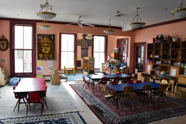 In a classroom with peach-colored walls, there are long, low tables with small chairs for children. The walls feature African tapestries, masks and other pieces of art as decoration.