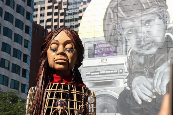 'Little Amal', a 12-foot-tall puppet of a little girl, stands in front of a mural of a little boy at Dewey Square in Boston. Her puppeteer, who is located inside the puppet's body, controls her facial expressions. The puppets eyes are closed as she faces the sun. 