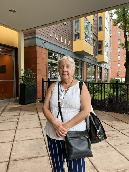 A woman in a white shirt and striped blue pants, carrying two black purses, stands in front of a building in Jamaica Plain.