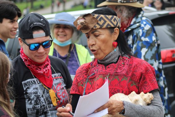 A woman wearing traditional indigenous attire, including a red top and brown headwear, speaks to another person in sunglasses and a black hat. She holds a paper in her left hand, pointing to something on the page with a pen in her right hand.