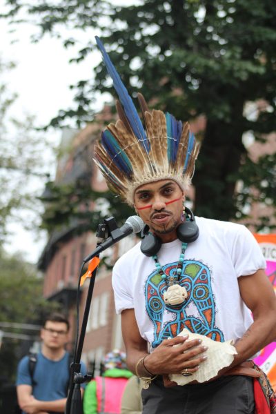 A young man stands in front of a microphone in Boston Common. He wears indigenous headwear from the Taíno people of Puerto Rico and a white t-shirt with a colorful indigenous print. He has traditional markings tattooed on his face. His nails are painted silver and he holds a conch shell.