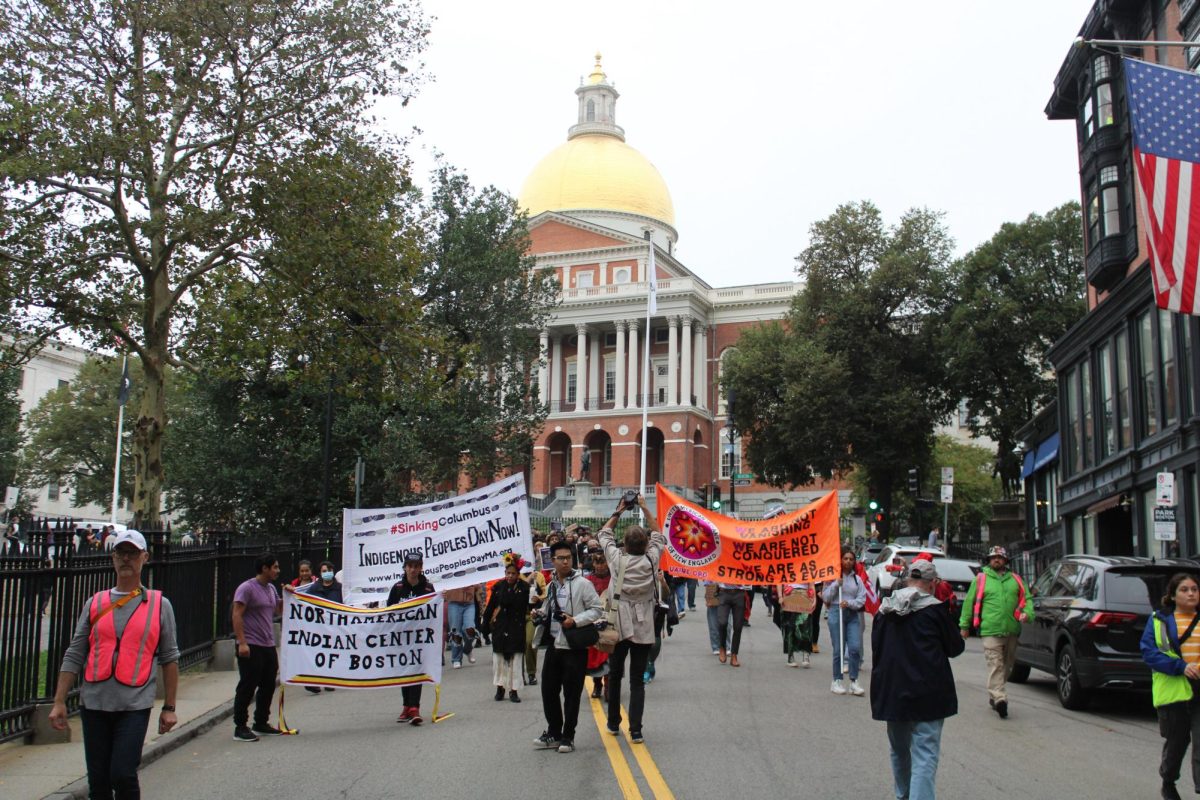 A crowd of people holding large banners and signs walk downhill, away from the Massachusetts State House in Beacon Hill. The people are marching for Indigenous Peoples Day. It is October; they are dressed warmly and the skies are gray.